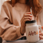 Ginger woman holding big cup of coffee. Stunning female model enjoying mulled wine at home.